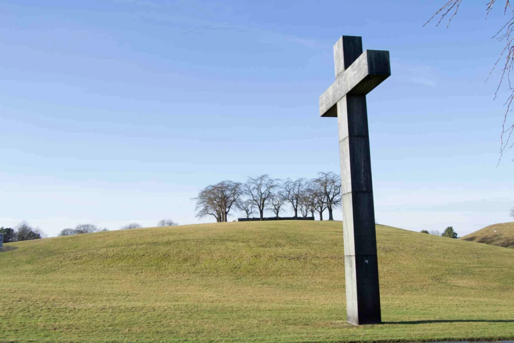 Ett stort stenkors står på en gräsbevuxen kulle med en grupp kala träd i bakgrunden under en klarblå himmel.