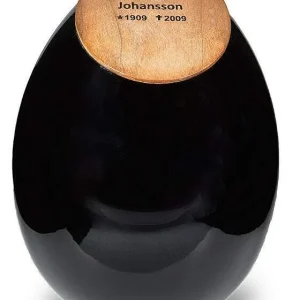 En svart, äggformad urna med ett lock av trä med namnet "Anna Johansson" ingraverat tillsammans med årtalen "1909" och "2009", vilket indikerar att det är en minnesurna.