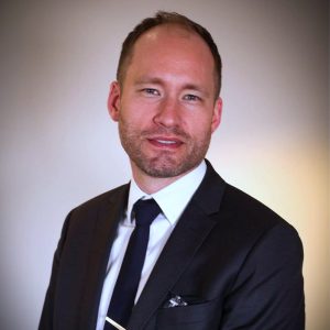 En man i mörk kostym, vit skjorta och svart slips står mot en vanlig, mjukt upplyst bakgrund och tittar in i kameran med ett litet leende.